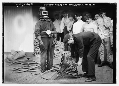 Black and white image of men standing around man in old fire fighter suit and air being pumped to him through a hose